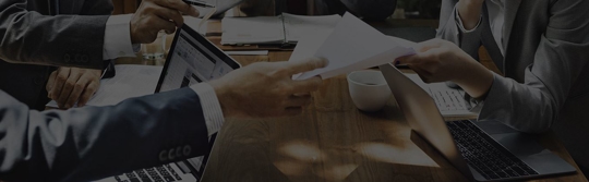 Hands exchanging documents over a conference table with laptops, representing workers’ comp application paperwork review.